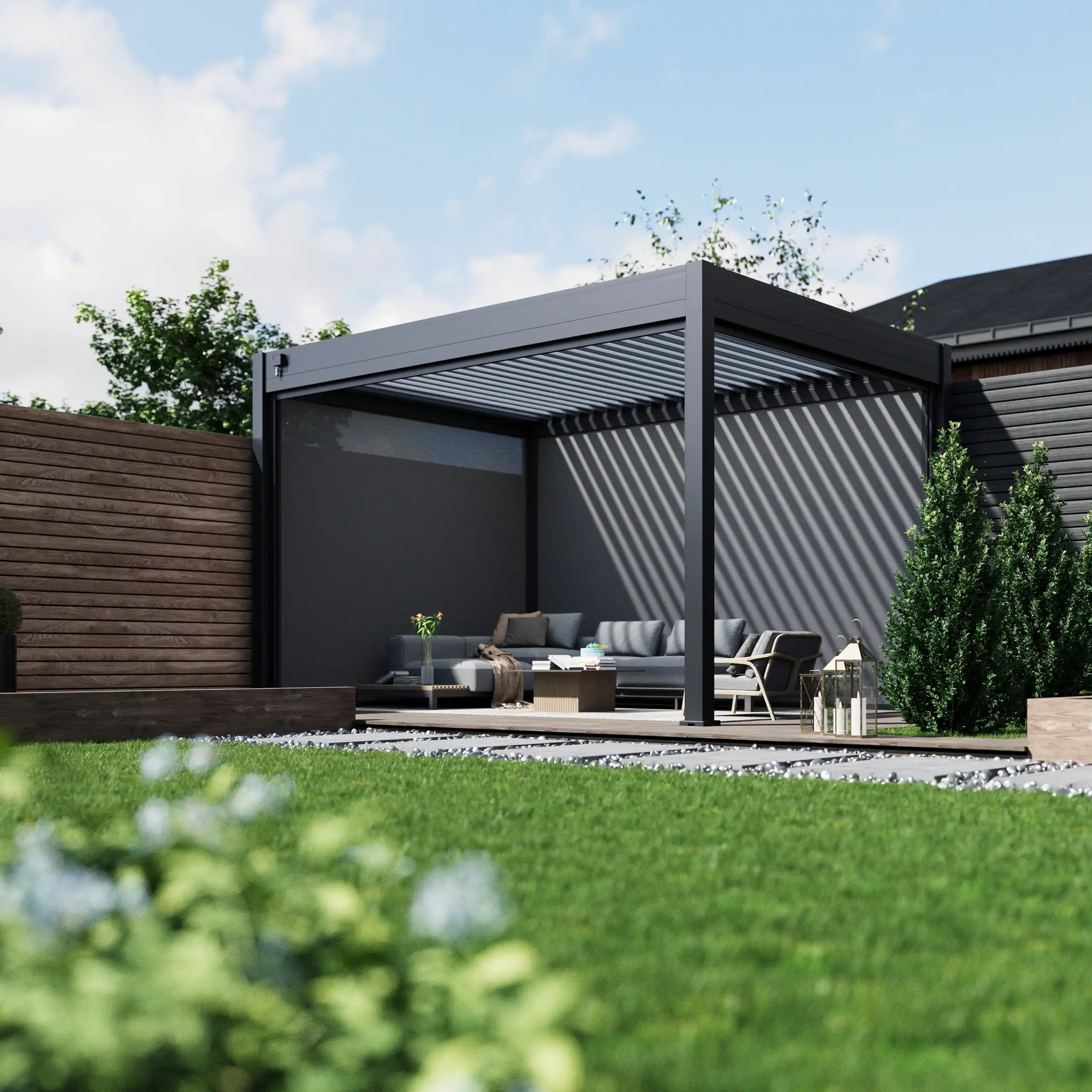 Freestanding dark grey aluminium pergola with an open louvred roof, located on a raised wooden deck with grey outdoor sofas, a coffee table, and neutral-toned cushions. Surrounded by lush green lawn, flower beds, and decorative lanterns, with timber fencing and tall hedging for added privacy.